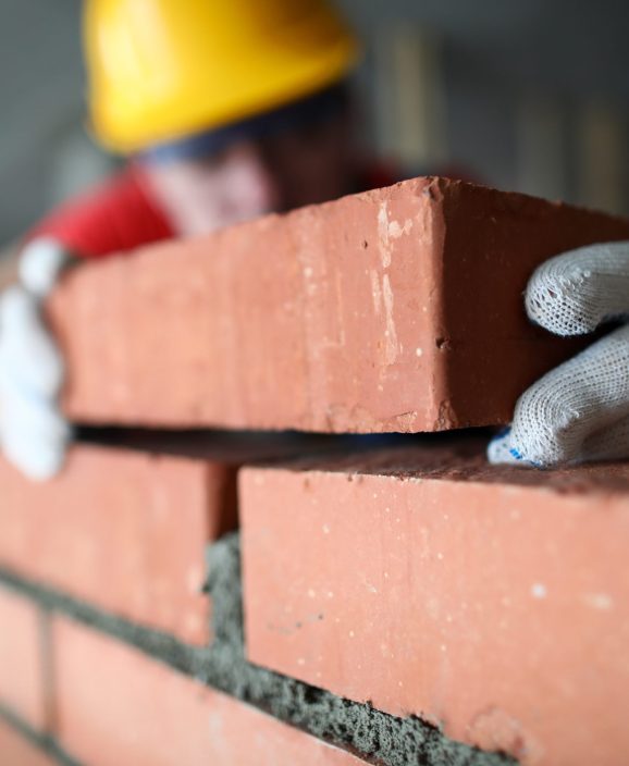 Close-up of professional construction worker laying bricks in industrial site. Builder in protective uniform. Man building wall with blocks. Renovation concept